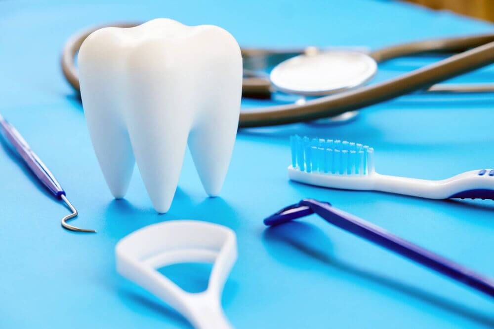 Large model tooth surrounded by a toothbrush, dental floss, mirror and dental instruments on a blue surface